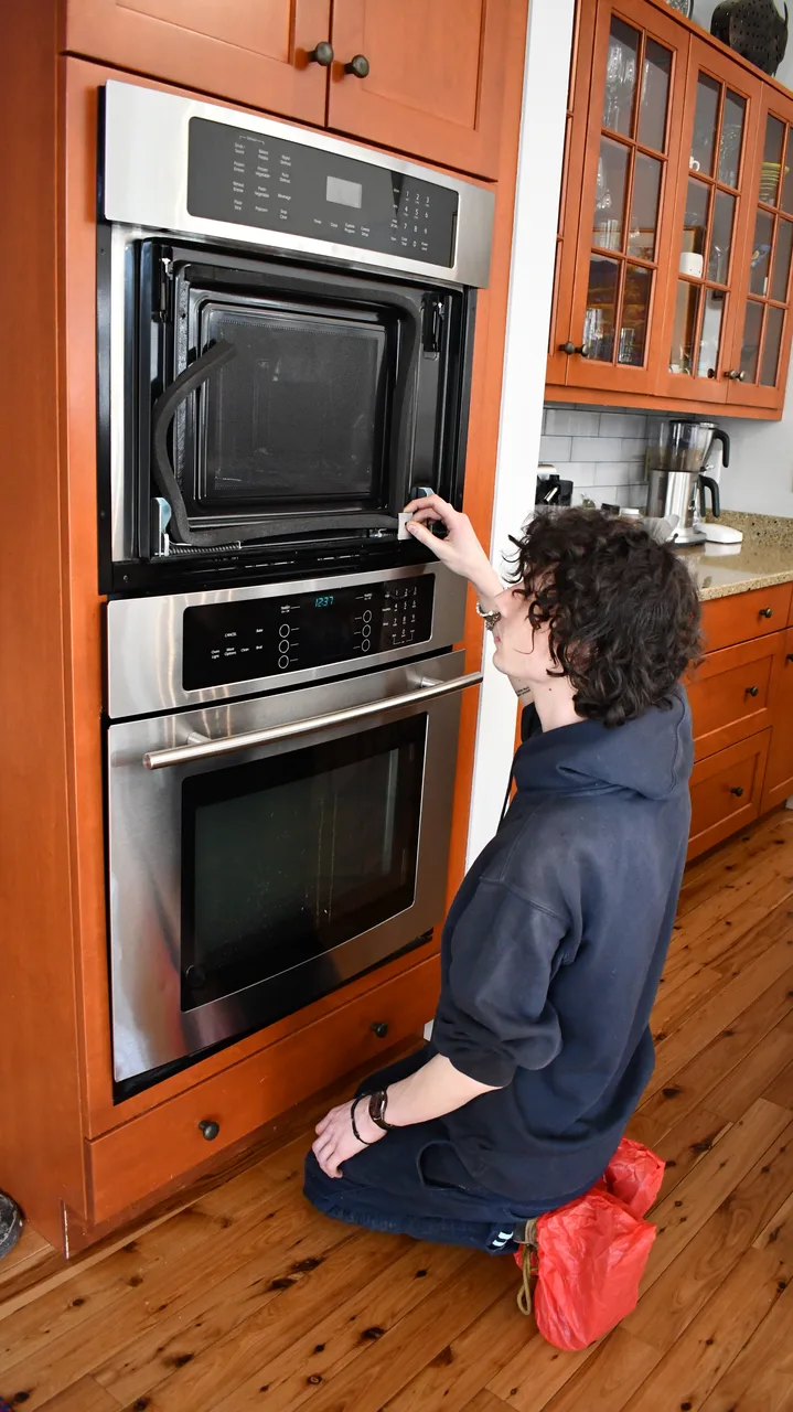 A technician kneeling on a wood floor to service a built-in wall oven and microwave