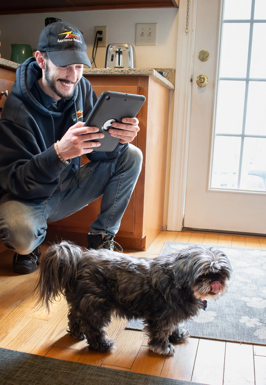 A technician in a cap crouching with a tablet in a home entryway beside a shaggy gray dog