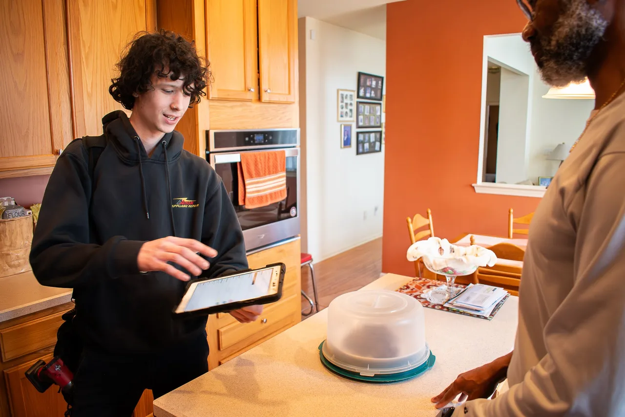 A technician reviewing a tablet at a customer's kitchen counter while the homeowner looks on